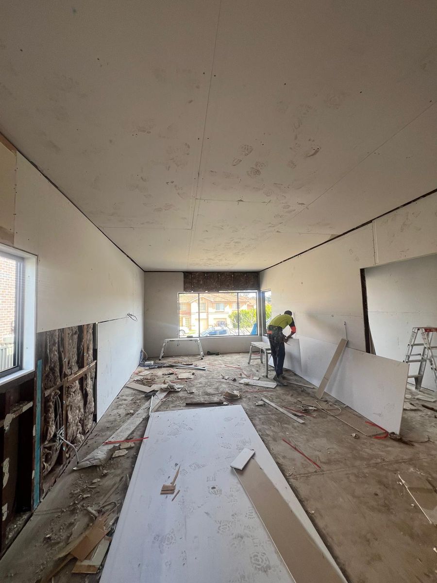 Plasterer in a hi-vis shirt positioning a new plasterboard sheet against a wall in a Sydney house, with the ceiling already sheeted and insulation batts visible in the open wall frame alongside