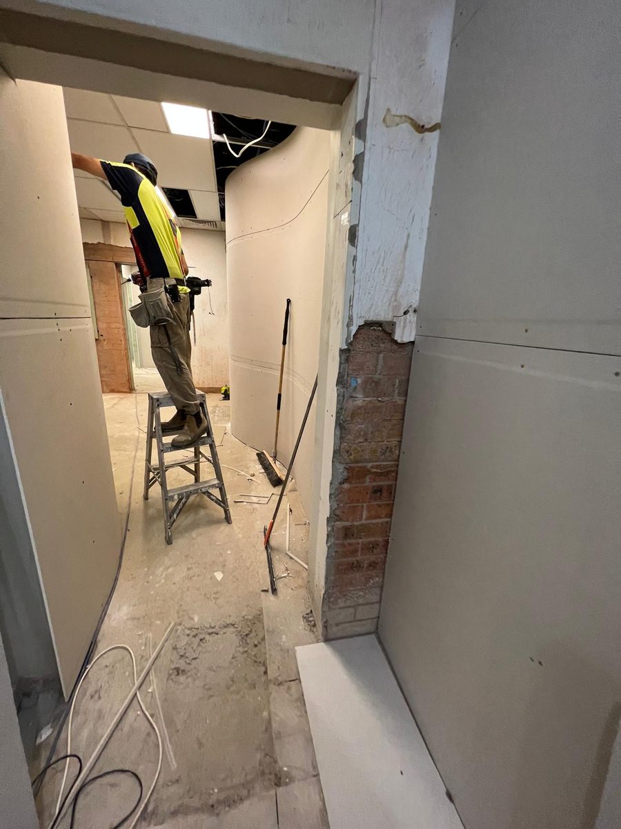 Plasterer on a ladder lining a narrow opening between rooms, with the brickwork exposed to one side