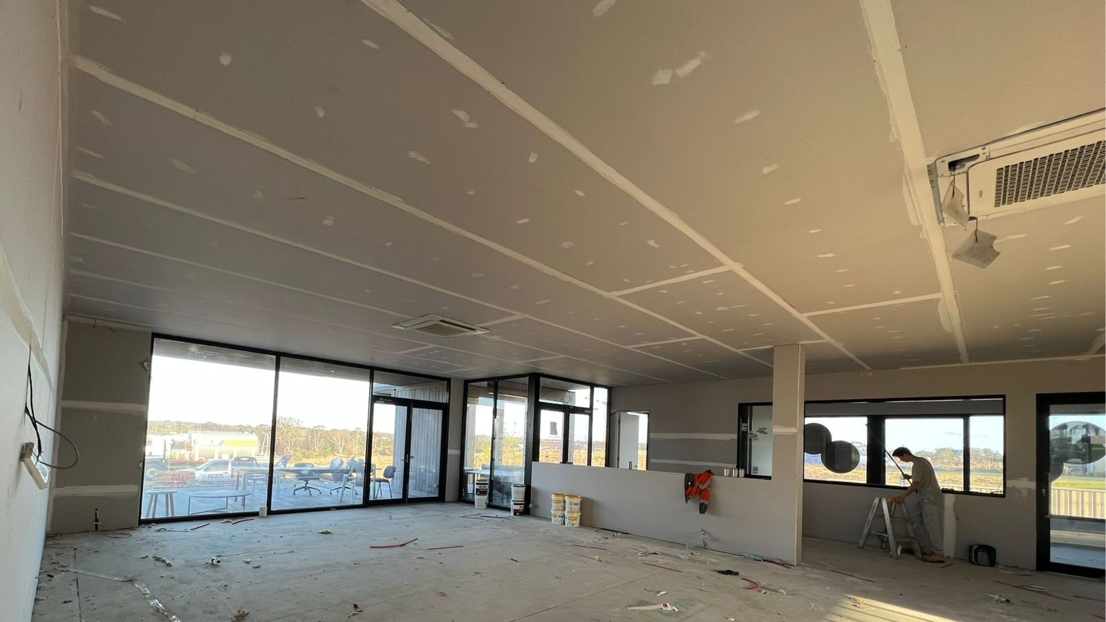 Commercial ceiling in a Sydney fit-out fully sheeted in plasterboard with every joint taped and topped, air-conditioning cassettes installed and a plasterer on a step ladder finishing a wall