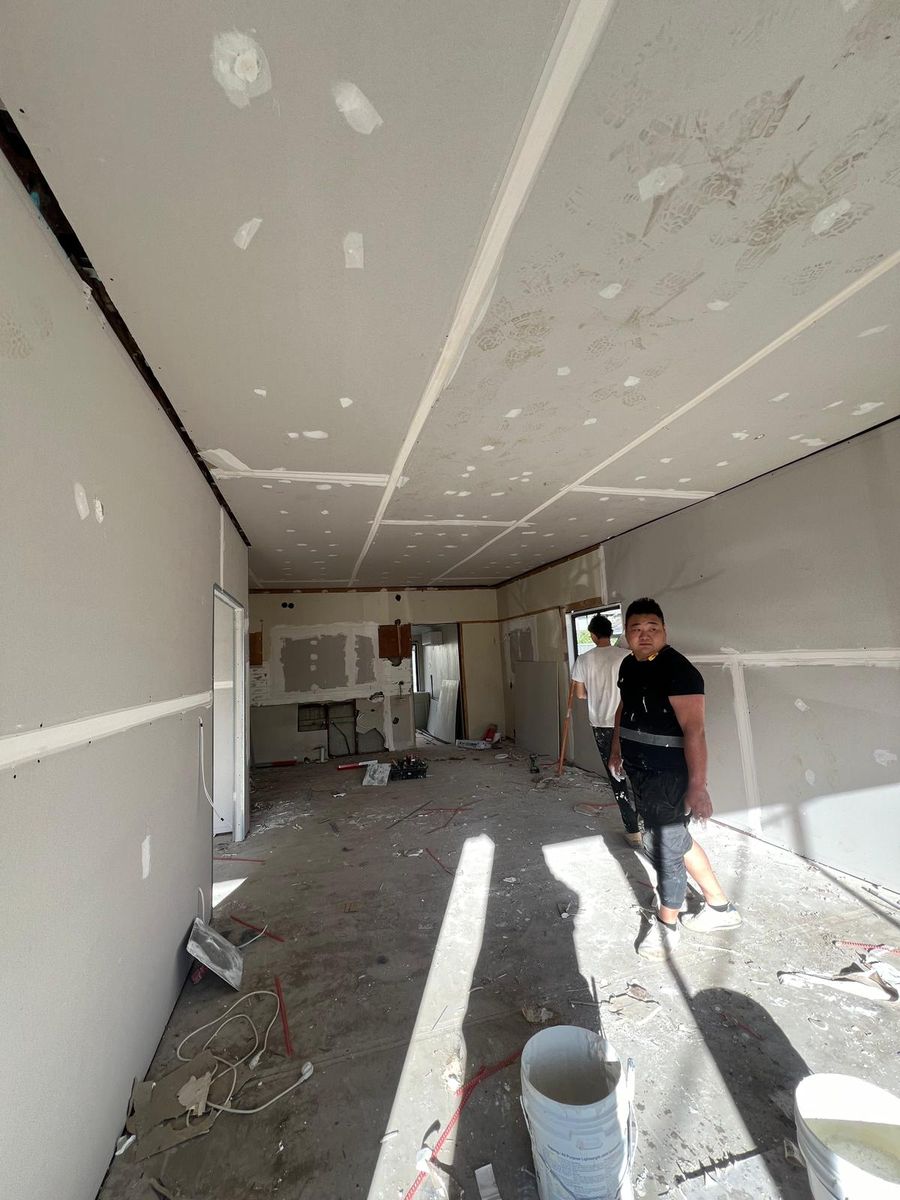 Two plasterers in a Sydney home renovation where the walls and ceilings have been sheeted and every joint taped and topped, with compound buckets and offcuts on the bare floor